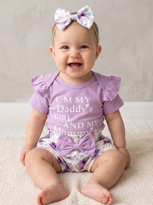 A smiling baby girl sitting on a textured cream blanket, wearing a lavender short-sleeve ruffle top with the text 'I'M MY Daddy's GIRL AND MY Mommy's' in white, matching floral-print diaper cover with a large purple bow, and a matching floral hair bow.