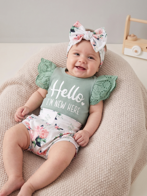 A baby lying on a textured beige beanbag chair, wearing a sage green ribbed top with lace flutter sleeves and the words 'Hello I'M NEW HERE' in white, matching floral bloomers, and a floral bow headband.
