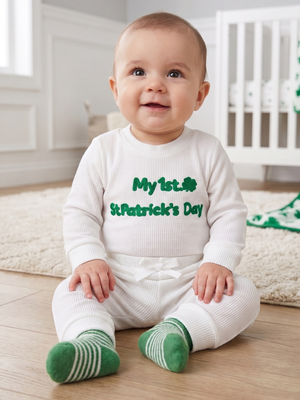 A baby sitting on a wooden floor in a nursery, wearing a white waffle-knit romper with green text reading 'My 1st St. Patrick's Day' and green-and-white striped socks.