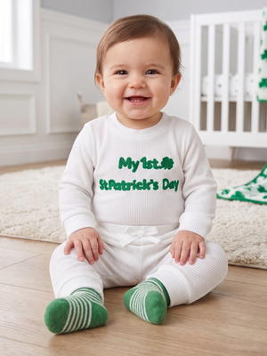 A smiling baby sitting on a wooden floor in a nursery, wearing a white waffle-knit one-piece outfit with green text reading 'My 1st St. Patrick's Day' and green-and-white striped socks.
