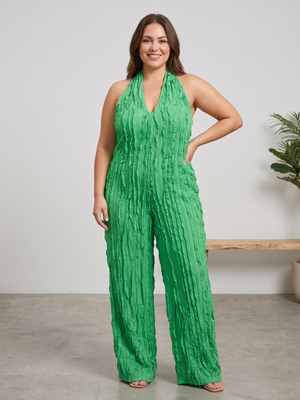 A woman modeling a bright green halter-neck jumpsuit with a crinkled texture and wide legs, standing in a minimalist studio setting with a light gray wall and concrete floor.