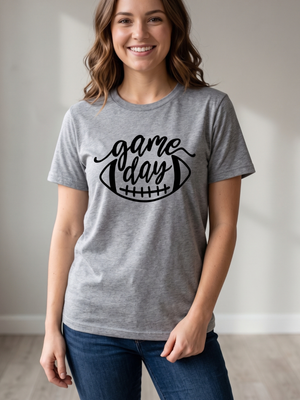 A woman wearing a heather gray t-shirt with a black graphic that reads 'game day' inside a football outline, paired with blue jeans in a brightly lit indoor setting.