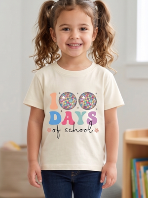 A young girl with curly brown hair in pigtails smiling while wearing a cream-colored t-shirt with a colorful '100 Days of School' graphic, paired with dark blue jeans in a brightly lit indoor room.