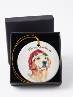 A round ceramic Christmas ornament featuring a Golden Retriever wearing a red Santa hat, presented in an open black gift box with a gold hanging cord. The ornament is printed with the text 'Merry Woolfmas'.