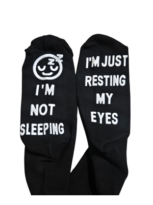 A pair of black crew socks with white embroidered text and graphics. One sock features a sleeping face emoji above the words 'I'M NOT SLEEPING', and the other features the text 'I'M JUST RESTING MY EYES'.