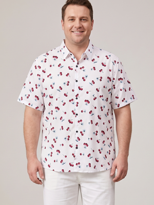 A man wearing a white short-sleeve button-up shirt with a red and blue cherry print, paired with white pants, standing against a light gray studio background.