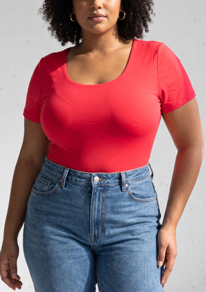 A woman wearing a bright red scoop-neck short-sleeve top and blue jeans, shown from the shoulders down against a light gray textured wall.
