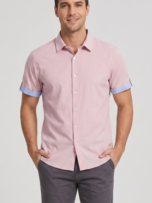 A man wearing a light pink short-sleeve button-down shirt with light blue and white striped rolled cuffs, paired with dark gray trousers, against a light gray studio background.