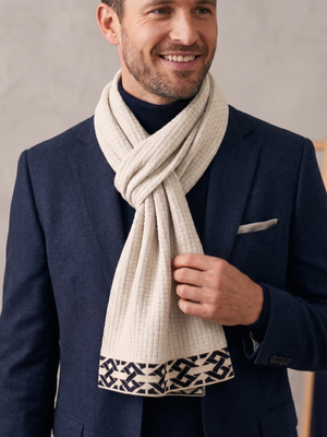 A man wearing a navy blue blazer and a cream-colored textured scarf with a black geometric patterned border, standing against a neutral gray background.