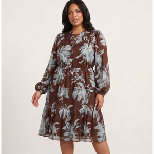 A woman modeling a brown long-sleeve midi dress with a gray and white floral print, featuring a tiered skirt and a keyhole neckline with drawstrings, set against a plain light beige studio background.