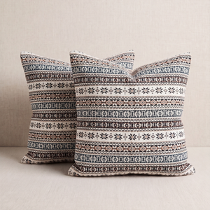 Two square throw pillows with a detailed Nordic/Fair Isle pattern in cream, brown, and navy blue, displayed on a light beige textured surface.