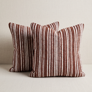 Two square throw pillows with a vertical rust-brown and off-white stripe pattern, arranged on a light beige textured surface against a matching neutral background.