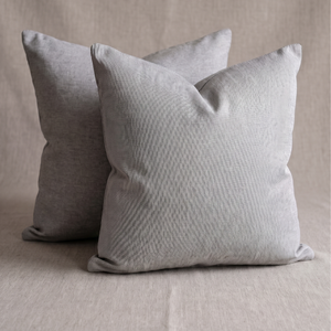 Two square throw pillows in a light gray textured fabric, with one pillow in front of the other on a neutral beige fabric surface.