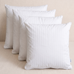 Four white rectangular pillow shams or decorative pillows arranged in a staggered row against a light beige textured background.