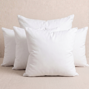 Four white square throw pillows arranged in a stack on a light beige textured surface against a matching beige background.