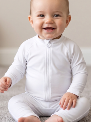 A smiling baby sitting cross-legged on a light carpet, wearing a white long-sleeve zip-front baby jumpsuit or one-piece.