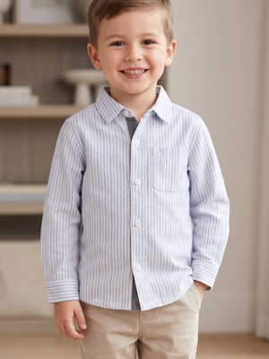 A young boy smiling and wearing a light blue and white vertically striped long-sleeve button-down shirt with beige pants, standing in a softly lit indoor room.