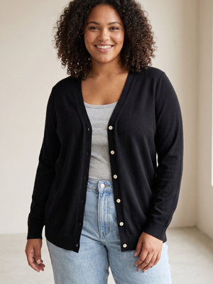 A woman with curly dark hair smiling, wearing a black button-up cardigan over a light gray tank top and light blue jeans, standing in a brightly lit indoor setting.