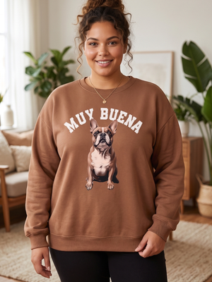 A woman wearing a brown crewneck sweatshirt with a graphic of a French Bulldog and the text 'MUY BUENA' in white, standing in a cozy, sunlit living room.