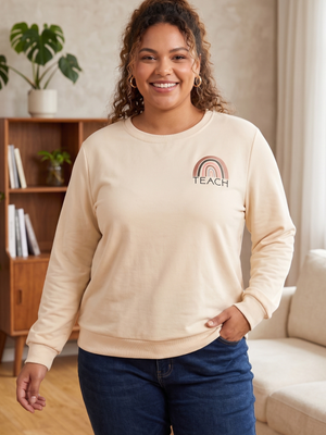 A woman wearing a cream-colored long-sleeve sweatshirt with a small rainbow graphic and the word 'TEACH' printed on the left chest, paired with dark blue jeans in a cozy indoor setting.