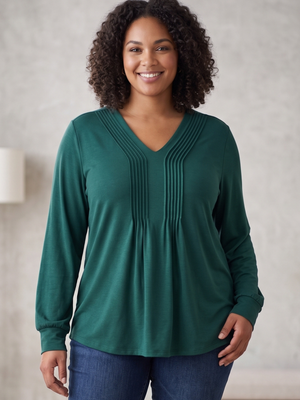 A woman with curly dark hair wearing a dark green long-sleeve V-neck top with vertical pleats on the chest, paired with blue jeans, standing against a neutral textured wall.