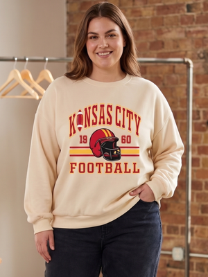 A woman wearing a cream-colored sweatshirt with a red and yellow 'Kansas City Football' graphic, paired with dark jeans, standing in front of a brick wall and clothing rack.