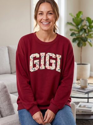 A woman sitting on a couch wearing a burgundy crewneck sweatshirt with the word 'GIGI' in a floral patterned font, paired with blue jeans in a bright living room setting.