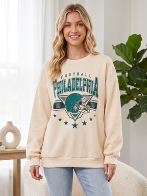 A woman wearing a cream-colored crewneck sweatshirt with a distressed teal and navy graphic that reads 'Football Philadelphia' with a football helmet, paired with light blue jeans in a bright indoor setting.