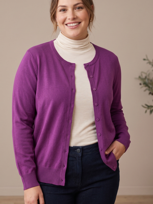 A woman wearing a purple button-front cardigan over a cream turtleneck sweater and dark blue jeans, standing against a neutral beige wall.