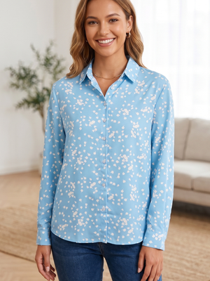 A woman wearing a light blue long-sleeve button-up shirt with a white floral print, paired with dark blue jeans, standing in a brightly lit living room.
