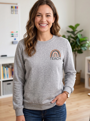 A smiling woman with wavy brown hair stands indoors wearing a heather gray crewneck sweatshirt with a small rainbow graphic and the word 'TEACH' on the left chest, paired with dark blue jeans.
