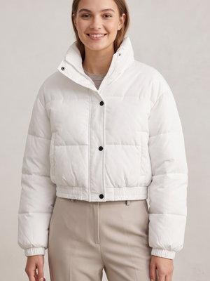 A woman wearing a cropped white puffer jacket with a high stand-up collar and black snap buttons, paired with beige trousers, against a light gray studio background.